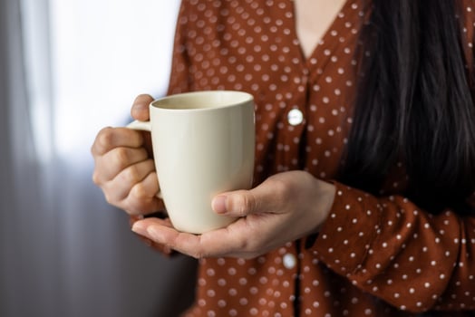 Close-up of asian woman holding mug, displaying comfort, warmth, and relaxation. Hands cradling neutral-colored cup, wearing red polka dot blouse, suggesting cozy, calm atmosphere. photo