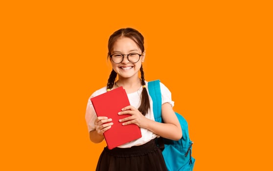 Finally, Back To School. Adorable Chinese Schoolgirl With Backpack Holding Book Standing On Yellow Background. Studio Shot photo