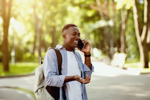 Happy black guy with backpack speaking on mobile phone at city park, copy space photo
