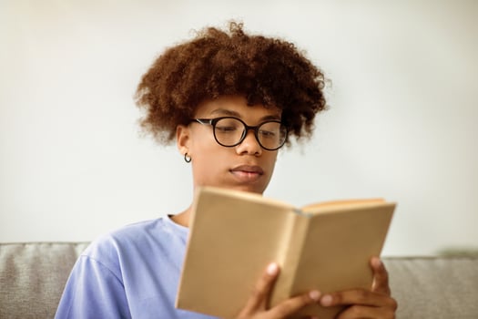 Focused African American teenager reading book, enjoying captivating story at home. Intelligent black teen student with textbook getting ready for test or exam, studying remotely indoors photo