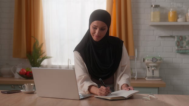 Smiling Islamic business woman freelancer Arabian girl student Muslim female lady businesswoman in hijab study work online internet in home kitchen typing laptop writing notes in notebook task write photo