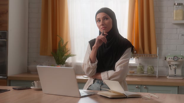 Pensive thinking thoughtful think Islamic Indian woman Muslim girl female in hijab homeowner freelancer business lady at home kitchen ponder come up find idea using laptop hold pen working computer photo