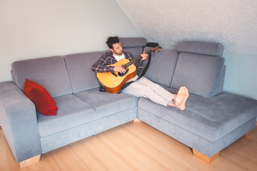 Barefoot musician sits comfortably on a gray couch, strumming an acoustic guitar in a cozy, relaxed atmosphere, lost in the soothing melodies. photo