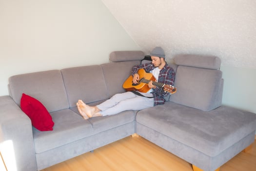 Barefoot musician lies on a gray couch, strumming an acoustic guitar in a cozy, relaxed atmosphere, lost in the soothing melodies photo