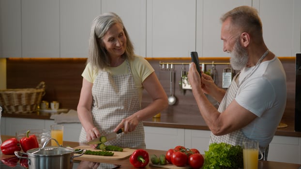 Retired old couple happy Caucasian family cooking in kitchen woman chef cutting cucumber chopping fresh healthy vegetables senior elderly man make photo on mobile phone cook on camera recording