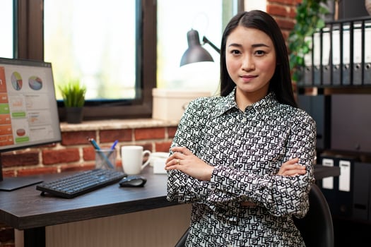 Closeup of businesswoman confidently posing with her arms crossed in modern workspace. Portrait of female entrepreneur sitting at office desk and looking at camera in brick wall room. photo