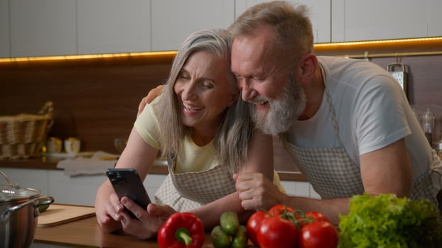 Laughing retired old gray haired Caucasian family love couple using mobile phone online order delivery food vegetables on kitchen table happy senior woman and man talking shopping cooking healthy meal photo