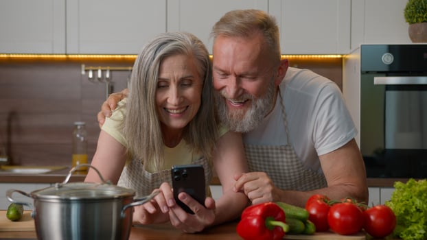 Caucasian married couple bonding family old mature woman man read cooking recipe on mobile phone senior 50s husband wife talking hugging shopping online food order with smartphone together in kitchen photo