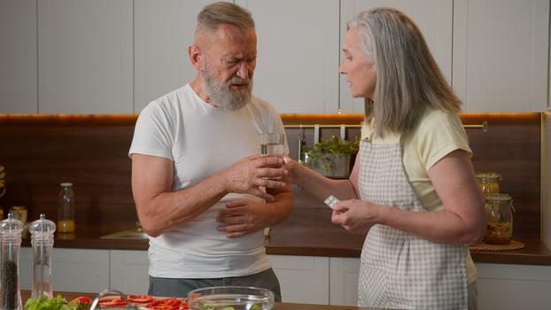Sick unhealthy old man in kitchen feeling pain in chest heart attack heartache bad health cardiac medical problem Caucasian family caring mature woman wife help husband give remedy tablet drink pill photo