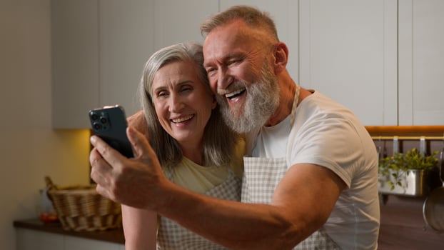 Mature retired Caucasian couple family old happy grandparents talk online call using mobile phone at home kitchen smiling senior elder woman wife and man husband waving hand to smartphone camera photo