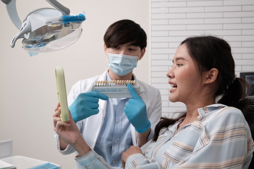 Male dentist explains color shades of tooth implant whitening to young Asian female patient, look at mirror in dental clinic, and do professional mouth hygiene healthcare work in a doctor's hospital. photo