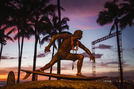 A bronze surfer statue adorned with flower leis stands against a vibrant sunset sky, with silhouetted palm trees adding to the tropical setting. photo