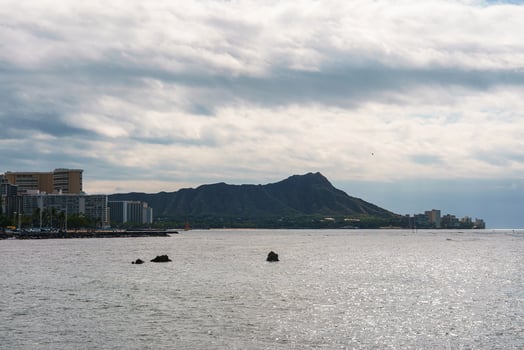 Diamond Head's ridged silhouette rises behind the Pacific Ocean, with Honolulu's modern skyline lining the coastline under a partly cloudy sky. photo