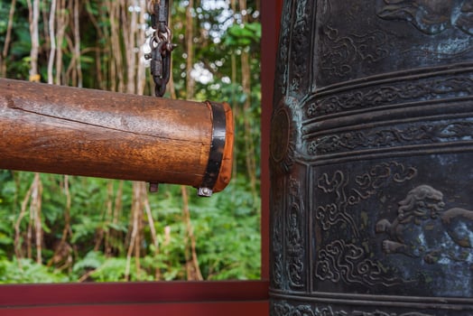 Traditional Japanese style bronze bell with intricate carvings and a wooden striker, set against lush green foliage at Byodo In Temple in Hawaii. photo