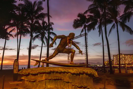 The bronze Duke Kahanamoku statue with flower leis stands against a vibrant sunset, surrounded by palm trees, Waikiki's skyline, and the beach. photo