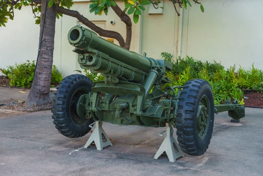 A green military artillery cannon rests on white stands outdoors, surrounded by tropical vegetation and a light colored building wall in Hawaii. photo