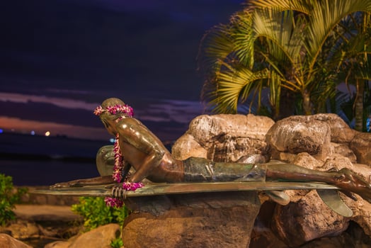 A bronze statue of a surfer on a surfboard adorned with flower leis, set against rocks, tropical plants, and an ocean backdrop under a darkening sky. photo