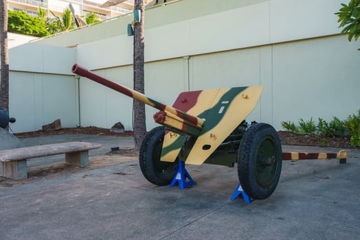 A vintage military artillery piece with camouflage paint on display outdoors, surrounded by palm trees, a concrete bench, and landscaped greenery. photo