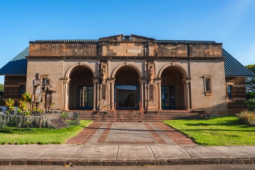 The Kauai Museum in Lihue, Hawaii features a classical facade with three arches, ornate columns, a statue, brick pathways, and a clear blue sky. photo