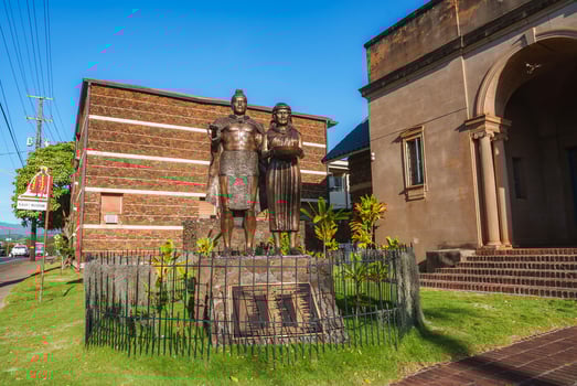 A bronze statue of a man and woman stands on a stone pedestal near a historic building with arched entrances, surrounded by tropical plants and a lawn. photo