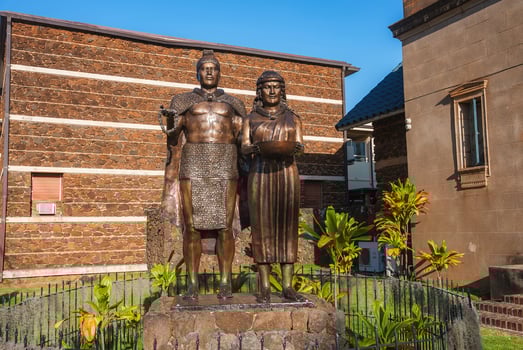 Bronze statue of a man in a cape and a woman holding a bowl, set on a stone pedestal with tropical plants and buildings in the background. photo