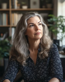 Thoughtful woman with gray hair sits indoors, surrounded by plants and books, in a patterned shirt. photo