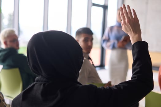 A Muslim businesswoman, dressed in professional attire and wearing a hijab, raises her hand during a business meeting to ask a question to the educator leading the session photo