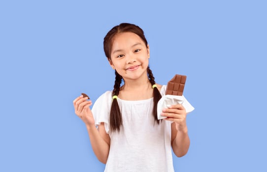 Chinese Little Girl Eating Chocolate Posing Over Purple Background In Studio, Smiling To Camera. Sweet Tooth Concept photo