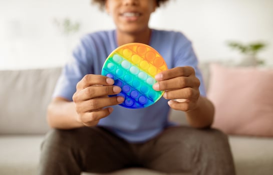 Cropped view of Afro teen guy playing with antistress POP IT toy indoors, closeup of hands. Unrecognizable black youth using sensory plaything at home. Stress relief, relax concept photo