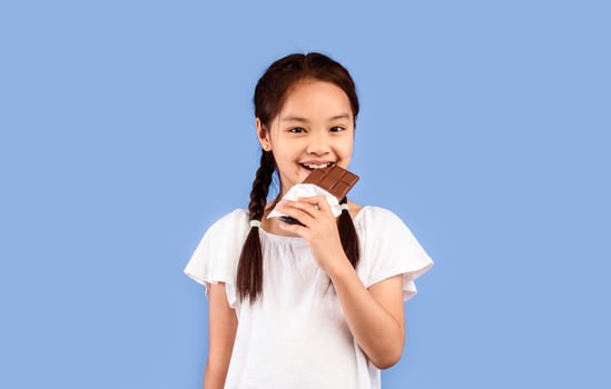 Sweet Tooth. Little Asian Girl Eating Chocolate Smiling To Camera Standing On Purple Background. Studio Shot photo