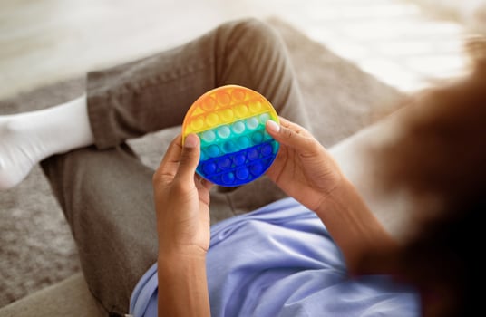 Cropped view of black teenager playing with antistress POP IT toy, sitting on sofa at home, closeup. Unrecognizable African American youth enjoying antistress sensory plaything, relaxing indoors photo