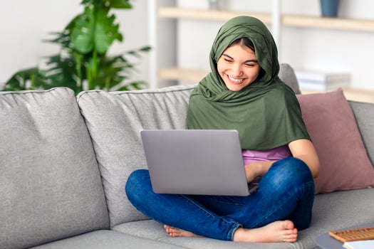 Remote learning concept. Happy Muslim teen girl in hijab studying online, sitting on sofa with laptop at home. Indian teenager in Islamic headscarf using PC, doing assignment, communicating to teacher photo