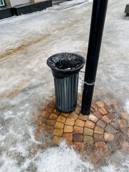 On a quiet, snowcovered sidewalk, a solitary trash can is placed next to a tall pole, which proudly stands amidst the serene winter landscape, adding a touch of urban charm photo