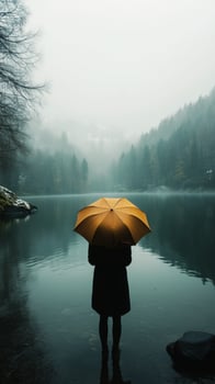 Person with yellow umbrella stands by tranquil lake in misty forest during early morning. photo