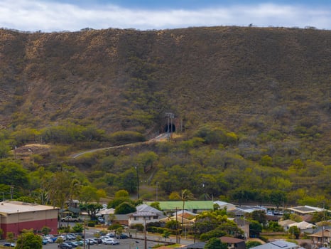 A hillside with a tunnel entrance surrounded by greenery, power lines, and a residential area with houses, parking lots, and larger buildings on Oahu. photo