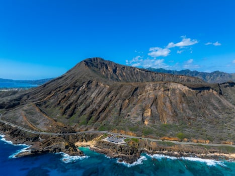 Aerial view of Diamond Head Crater on Oahu Island, Hawaii, with rugged slopes, a coastal road, turquoise waters, rocky cliffs, and a clear blue sky. photo