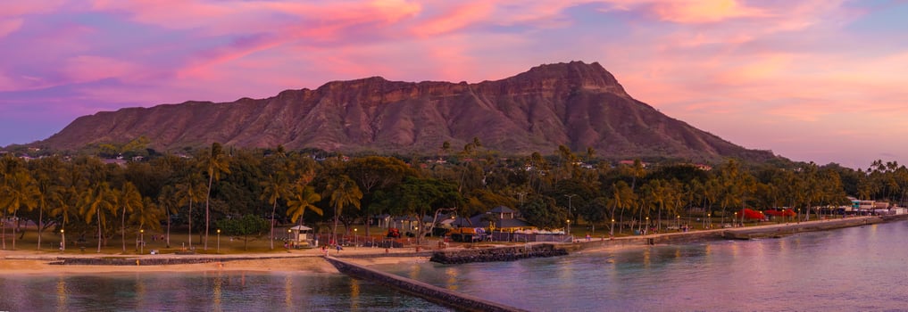 View of Diamond Head Crater during sunset with a serene beach, palm trees, walking path, and calm ocean reflecting pink and purple sky hues. photo