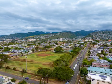 Aerial view of a residential area on Oahu island, Hawaii, featuring a central park with a baseball field, a main road, and lush mountains in the background. photo