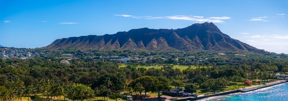 Diamond Head Crater with rugged ridges, lush greenery, palm trees, and a coastal area bordered by a seawall, under a bright blue sky with clouds. photo