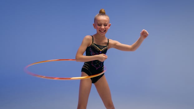 Caucasian little girl child kid daughter schoolgirl gymnast athlete dancer ballerina acrobat stand in split position flexibility artistic gymnastics spinning hoop sport event studio blue background photo