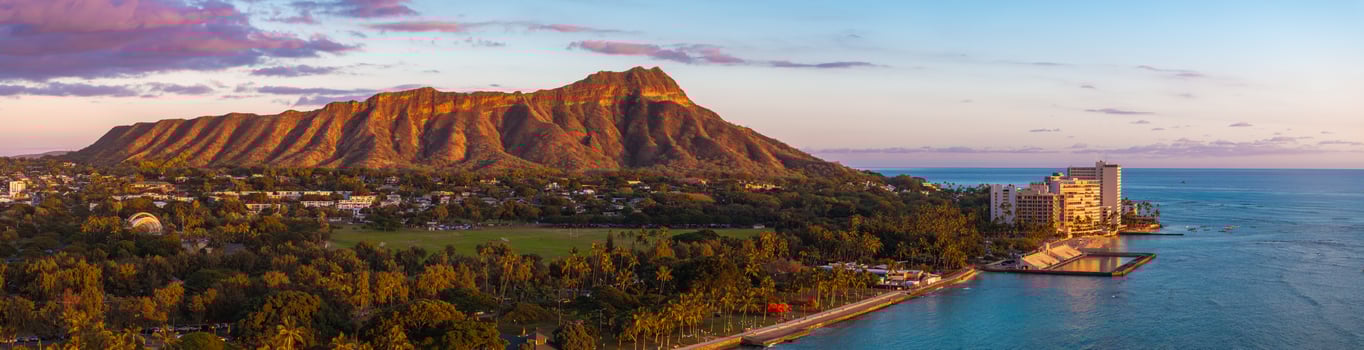 Diamond Head Crater on Oahu Island, Hawaii, at sunset with golden light, lush greenery, urban areas, and turquoise Pacific Ocean waters visible. photo