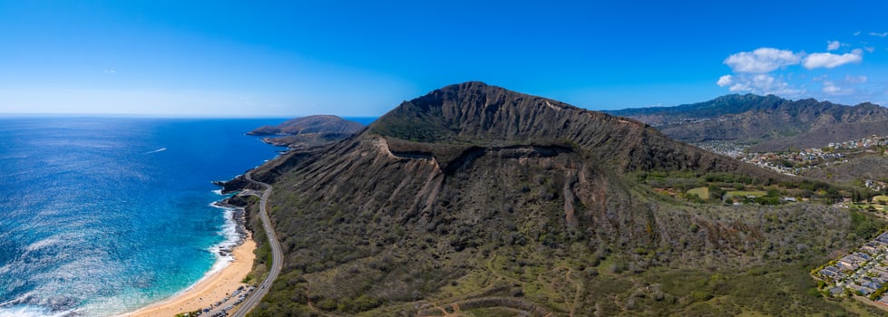 Aerial view of Diamond Head Crater, sandy beach with turquoise waters, winding road, residential area, verdant mountains, and clear blue sky. photo