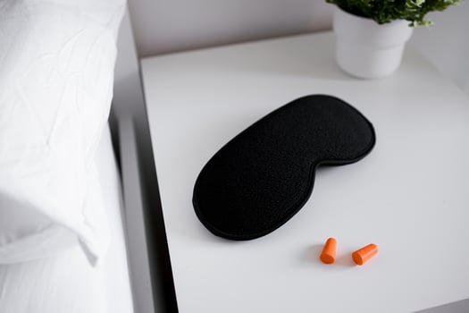 Black sleep mask and orange earplugs on white nightstand with green plant photo