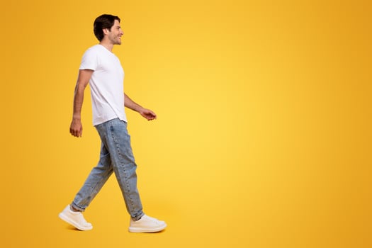 Full length body size profile side view of happy excited bearded guy in casual white t-shirt and jeans walking looking aside at free empty space isolated over orange color background, panoramic banner photo