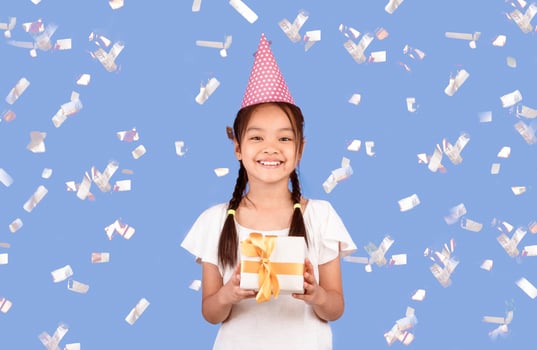Kid's Birthday Party. Chinese Girl Holding B-Day Gift Box Standing Under Falling Confetti On Purple Background. Studio Shot photo