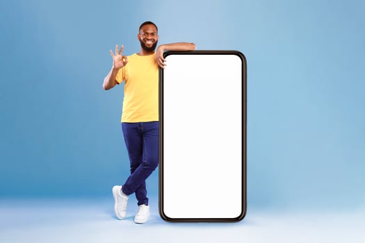 Positive African American guy showing okay gesture next to huge smartphone on blue studio background, empty space for mobile app design on screen. Website or advertisement mockup photo