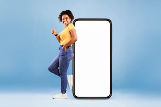 Pretty African American lady standing near big smartphone with blank screen, gesturing thumb up on blue background, mockup. Young woman recommending new mobile app, advertising website, free space photo