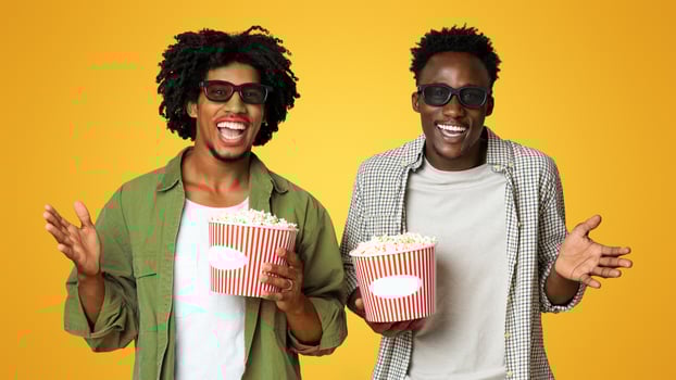 Movie Day. Two happy black male friends in 3d glasses with popcorn buckets posing over beige background, excited cheerful young african american mates having fun in cinema, enjoying watching film photo