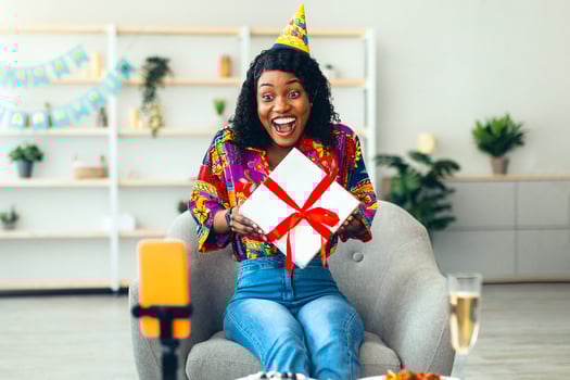 Online Birthday Party. Excited African Woman Holding Wrapped Gift Box Having Call By Phone Receiving B-Day Presents Celebrating Sitting In Living Room At Home. Distant Holiday Celebration photo