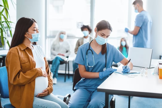 flu Vaccination Concept. Woman Doctor In Face Mask Taking Notes Talking With Pregnant Woman Patient Before Coronavirus Vaccine Injection Sitting In Hospital Room, Young Asian Lady Hugging Belly photo